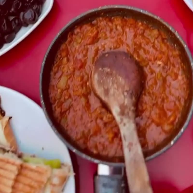 Dükkanda çekilmiş fotoğraf: kırmızı masada tahta kaşıklı menemen tavası.